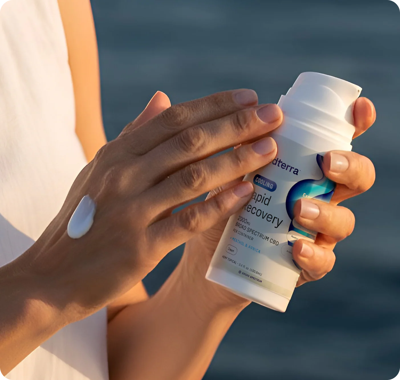 Person applying Rapid Recovery cooling CBD cream from a white pump bottle by the ocean