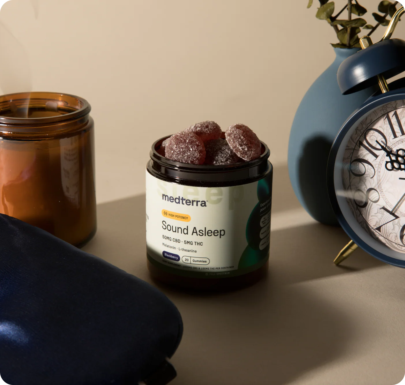 Front of Sound Asleep jar with open sugar-coated gummies and label 50mg CBD 5mg THC, staged with alarm clock and sleep mask